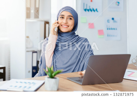 Business Communication. Joyful Arabic Female Employee Talking On Cellphone And Using Laptop While Working In Modern Office Business Communication. Joyful Arabic Female Employee Talking On Cellphone And Using Laptop While Working In Modern Office 132871975