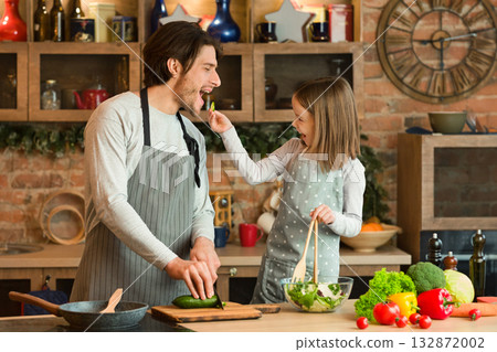 Happy Young Family Father And Daughter Having Fun While Cooking In Kitchen Together, Little Girl Feeding Dad With Vegetables While Preparing Healthy Food At Home, Making Lunch Or Dinner, Free Space Happy Young Family Father And Daughter Having Fun While Cooking In Kitchen Together, Little Girl Feeding Dad With Vegetables While Preparing Healthy Food At Home, Making Lunch Or Dinner, Free Space 132872002