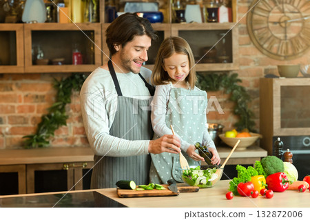 Cheerful young dad and little daughter preparing healthy salad in kitchen together, cooking lunch ant home, free space 132872006