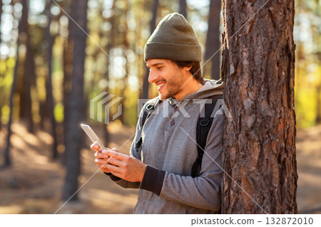 Happy young man with backpack chatting with friends on phone in forest, copy space 132872010