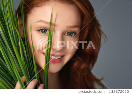 Beauty Portrait Of Red-Haired Young Woman Posing With Green Plant Looking At Camera Through Leaves Standing Over Gray Background, Studio Shot. Facial Skin Care And Cosmetics 132872020