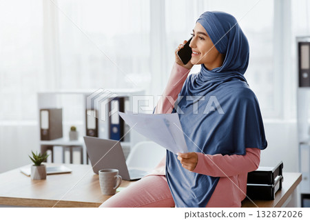Millennial muslim businesswoman holding papers and talking on cellphone in office, discussing contract conditions with partner, copy space 132872026
