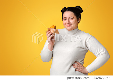 Beautiful plump woman posing with burger in hand over yellow background and smiling at camera, empty space Beautiful plump woman posing with burger in hand over yellow background and smiling at camera, empty space 132872058