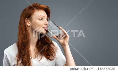 Healthy Hydration. Red-Haired Millennial Woman Drinking Water From Glass Standing Over Gray Studio Background, Wearing Casual T-Shirt. Side View Shot. Stay Hydrated Concept Healthy Hydration. Red-Haired Millennial Woman Drinking Water From Glass Standing Over Gray Studio Background, Wearing Casual T-Shirt. Side View Shot. Stay Hydrated Concept 132872080