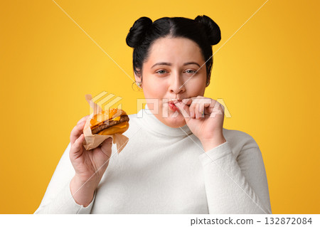 Young obese girl eating burger and licking fingers, enjoying fast food, high calorie nutrition Young obese girl eating burger and licking fingers, enjoying fast food, high calorie nutrition 132872084