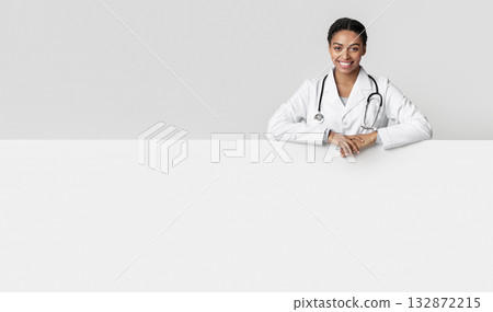 Medical ad. Millennial afro female doctor leaning on big blank advertising board with free space for your text or offer, posing over light background Medical ad. Millennial afro female doctor leaning on big blank advertising board with free space for your text or offer, posing over light background 132872215