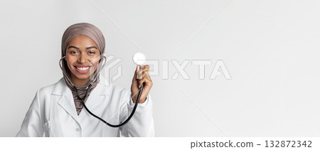 Portraif of young black muslim female doctor in hijab with stethoscope, posing over light background and smiling at camera, free space 132872342