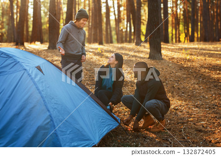 Cheerful multiracial backpackers setting camp in autumn forest, sun flare, free space 132872405