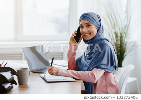 Portrait Of Arabic Female Secretary Talking On Cellphone And Taking Notes While Working On Laptop In Modern Office, Side View Portrait Of Arabic Female Secretary Talking On Cellphone And Taking Notes While Working On Laptop In Modern Office, Side View 132872406