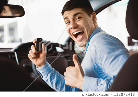 New Owner. Excited Guy Holding Car Key And Gesturing Thumb Up Sitting In Auto In Dealership Store 132872418