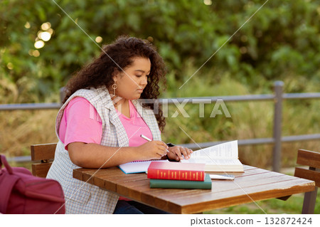 A student engaged in studying while seated at a wooden table outdoors. The individual writes in a notebook, with books beside them, immersed in their studies on a sunny day. A student engaged in studying while seated at a wooden table outdoors. The individual writes in a notebook, with books beside them, immersed in their studies on a sunny day. 132872424