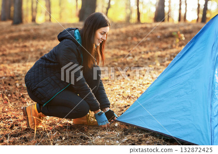 Happy adventurous girl setting camp in autumn forest, placing tent, empty space Happy adventurous girl setting camp in autumn forest, placing tent, empty space 132872425
