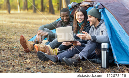 Happy international friends sitting in tent, using laptop and talking, forest background, copy space Happy international friends sitting in tent, using laptop and talking, forest background, copy space 132872456