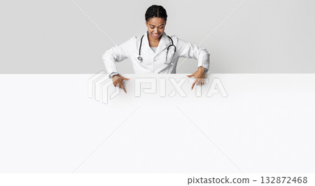 Medical offer. Afro female doctor looking down and pointing at blank white advertising board with empty space for text 132872468