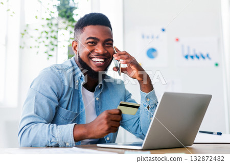 Shopping online. Cheerful african american guy talking on phone to internet shop manager, discussing details of his order while working in office. Shopping online. Cheerful african american guy talking on phone to internet shop manager, discussing details of his order while working in office. 132872482