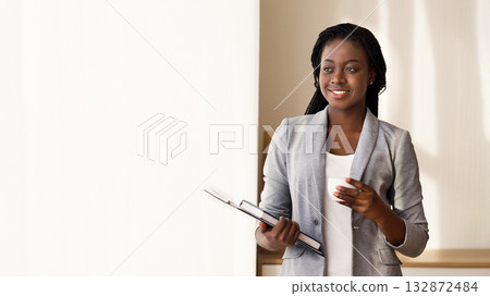 Pensive african secretary having coffee break near window in office, standing with clipboard, notebook and cup in hands, free space Pensive african secretary having coffee break near window in office, standing with clipboard, notebook and cup in hands, free space 132872484
