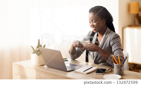 Coffee break. Successful black businesswoman holding cup with hot drink at workplace and looking at laptop screen, copy space Coffee break. Successful black businesswoman holding cup with hot drink at workplace and looking at laptop screen, copy space 132872509