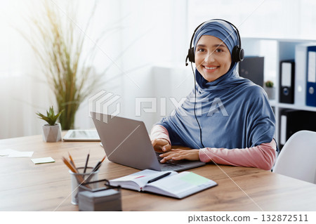 Jobs For Muslim Women. Portrait of happy arab woman call center operator wearing hijab and headset, working on laptop in office and looking at camera Jobs For Muslim Women. Portrait of happy arab woman call center operator wearing hijab and headset, working on laptop in office and looking at camera 132872511