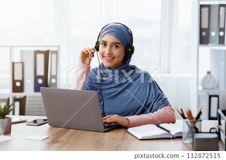 Online Conference Concept. Beautiful muslim female manager wearing headset having video call with partners, sitting at desk in office, free space Online Conference Concept. Beautiful muslim female manager wearing headset having video call with partners, sitting at desk in office, free space 132872515