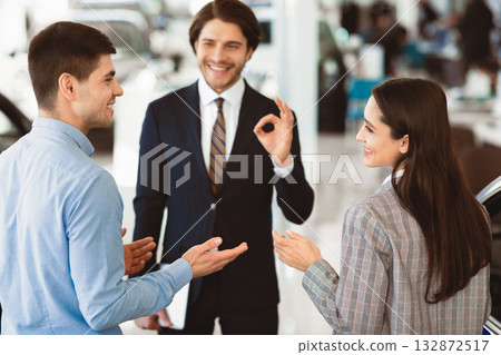 Nice Choice. Happy young couple talking about new car purchase, auto sales manager showing okay sign in the background Nice Choice. Happy young couple talking about new car purchase, auto sales manager showing okay sign in the background 132872517