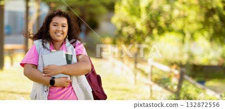 A young student wearing a pink shirt smiles as she walks through a vibrant park, holding notebooks close to her chest on a sunny day. The scene is filled with lush greenery. 132872556