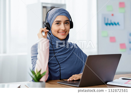 Call Center Operator. Portrait of beautiful arabic woman in hijab wearing headset, working at laptop in modern office, consulting clients. Call Center Operator. Portrait of beautiful arabic woman in hijab wearing headset, working at laptop in modern office, consulting clients. 132872581
