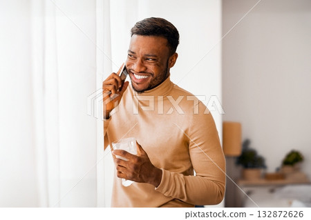 Phone Call. Joyful Afro Guy Talking On Cellphone Having Coffee Standing Next To Window At Home. Copy Space 132872626