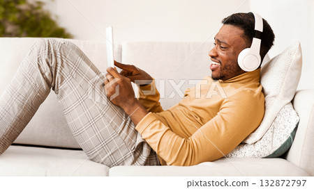 Weekend Concept. Happy African American Guy In Headphones Using Tablet Computer Listening To Music Relaxing Lying On Couch Indoor Weekend Concept. Happy African American Guy In Headphones Using Tablet Computer Listening To Music Relaxing Lying On Couch Indoor 132872797