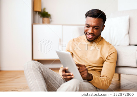 Afro Man Using Digital Tablet Reading Book Online Sitting On Floor At Home. Selective Focus Afro Man Using Digital Tablet Reading Book Online Sitting On Floor At Home. Selective Focus 132872800