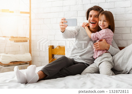 Happy preschool girl and father taking selfie on smartphone, laying on bed in morning, free space Happy preschool girl and father taking selfie on smartphone, laying on bed in morning, free space 132872811