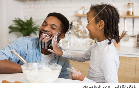 Happy african dad and daughter having fun while cooking pastry at home, girl slapping father face with flour palm Happy african dad and daughter having fun while cooking pastry at home, girl slapping father face with flour palm 132872931