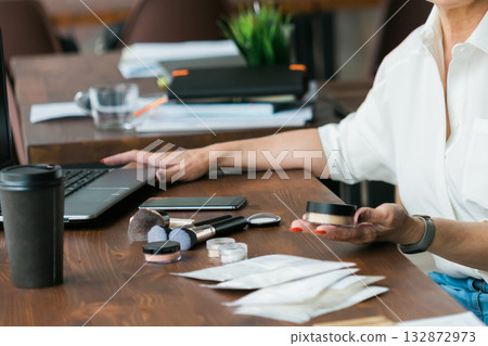 Woman holding cosmetic jar with mineral powderat wooden desk with laptop and coffee cup. Balance between work and self-care, illustrating modern lifestyle and routine. Woman holding cosmetic jar with mineral powderat wooden desk with laptop and coffee cup. Balance between work and self-care, illustrating modern lifestyle and routine. 132872973