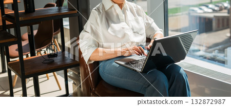 Woman typing on laptop near window in bright cafe interior. Banner with copy space and empty place for advertising. Focus, creativity, and productive work atmosphere in modern lifestyle. 132872987