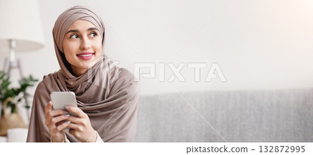 Portrait of pensive arabic girl in hijab with smartphone in hands sitting on sofa at home and looking away 132872995
