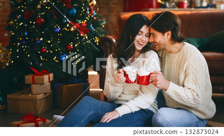 Young couple in love having xmas cocoa and bonding next to Christmas tree at home, free space Young couple in love having xmas cocoa and bonding next to Christmas tree at home, free space 132873024