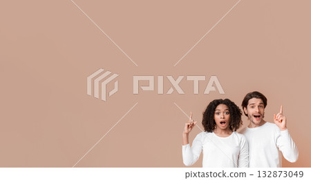 Great Offer. Interracial couple pointing up at copy space above their heads with fingers, posing over yellow background and looking at camera Great Offer. Interracial couple pointing up at copy space above their heads with fingers, posing over yellow background and looking at camera 132873049