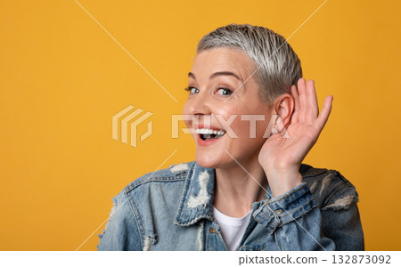 Gossip Concept. Curious mature lady holding hand near ear, trying to hear information, standing on yellow studio background, copy space 132873092