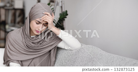 Unhappy muslim woman in headscarf sitting on sofa and touching head in despair, feeling depressed and lonely, having headache Unhappy muslim woman in headscarf sitting on sofa and touching head in despair, feeling depressed and lonely, having headache 132873125