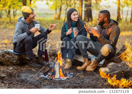Camping time for mixed race group of friends sitting beside fireplace and frying sausages in autumn forest in sun lights 132873129