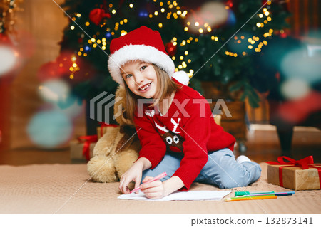 Xmas smiling girl sitting next to festive tree and drawing, looking at camera, copy space Xmas smiling girl sitting next to festive tree and drawing, looking at camera, copy space 132873141