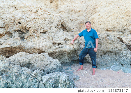 Portrait of an adult man in a blue shirt standing near a rock on the beach Portrait of an adult man in a blue shirt standing near a rock on the beach 132873147