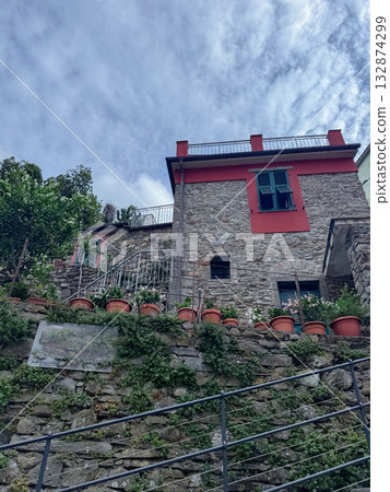 Stone house with red accents and green windows nestled on a hilltop. Decorated with numerous potted plants, it stands against a partly cloudy sky, showcasing rustic architecture. 132874299