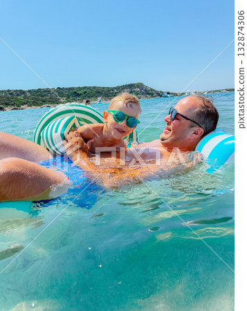 Happy boy and his dad playing in the water on a summer day. Family time at the beach, fun in clear sea water, father and child togetherness, vacation lifestyle, parenting and love concept. 132874306