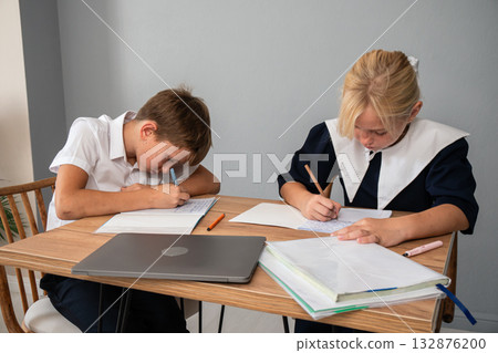 Children School Homework Studying - Two children in school uniforms sit at a table writing in notebooks while doing their homework. 132876200