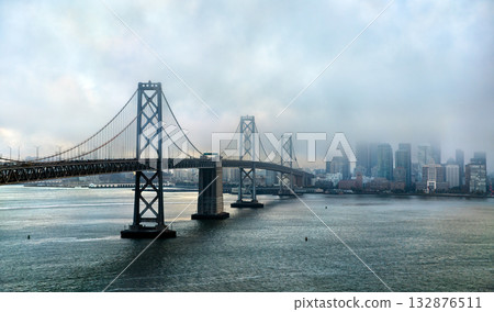 San Francisco-Oakland Bay Bridge on a moody, foggy day. The city skyline is partially hidden by low clouds, and the bay water is calm in the foreground. Represents travel and urban atmosphere 132876511