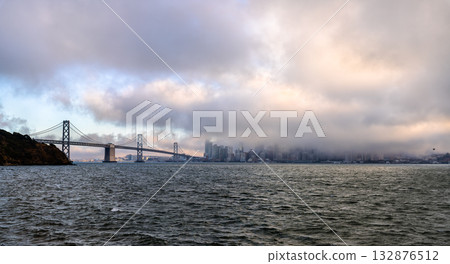 A moody, panoramic view of the San Francisco skyline mostly hidden by thick, low fog. The Bay Bridge is visible on the left, spanning the choppy bay water under a dramatic, cloudy sky A moody, panoramic view of the San Francisco skyline mostly hidden by thick, low fog. The Bay Bridge is visible on the left, spanning the choppy bay water under a dramatic, cloudy sky 132876512