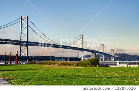 The San Francisco-Oakland Bay Bridge on a sunny day, viewed from a waterfront park with vibrant green grass. This image represents travel, infrastructure, and California landmarks The San Francisco-Oakland Bay Bridge on a sunny day, viewed from a waterfront park with vibrant green grass. This image represents travel, infrastructure, and California landmarks 132876520