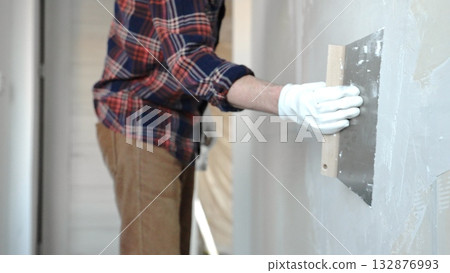 Unknown male construction worker wearing protective gloves smoothing plaster on interior wall during home renovation, applying material with professional trowel technique, close-up Unknown male construction worker wearing protective gloves smoothing plaster on interior wall during home renovation, applying material with professional trowel technique, close-up 132876993