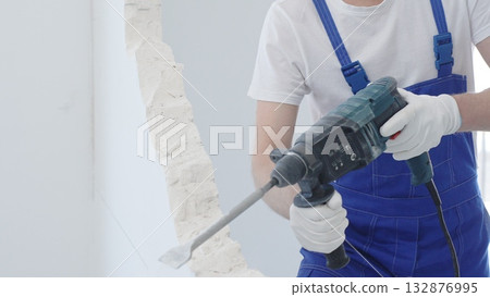 Male construction worker demolishing white wall with rotary hammer drill, wearing blue overalls and protective white gloves, closeup view 132876995