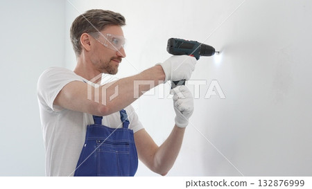 Middle aged man wearing blue overall and protective glasses, is drilling hole in white wall, wearing protective gear during home improvement project with cordless power tool 132876999